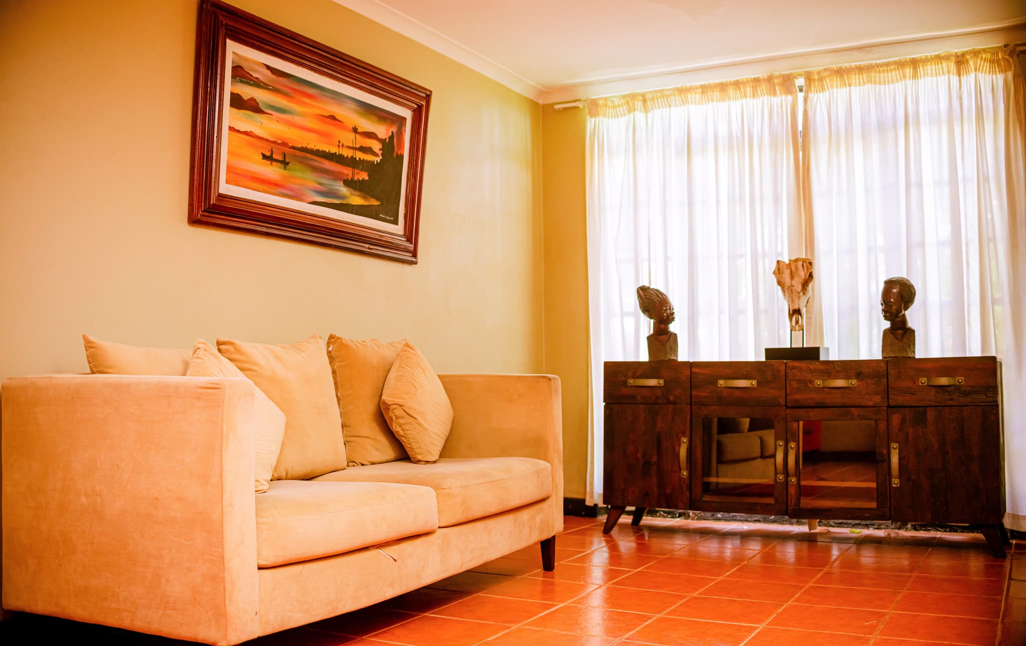 Cozy hotel lounge with beige sofa, local artwork, and wooden cabinet with African sculptures.